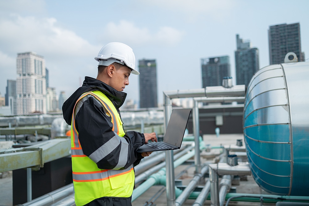 A focused engineer in a reflective vest and hard hat uses a laptop on an urban industrial rooftop, inspecting equipment.