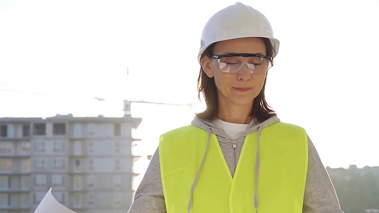 Female architect or engineer wearing safety hard hat and vest holding blueprint while inspecting a construction site at sunrise, front view. Architecture concept.